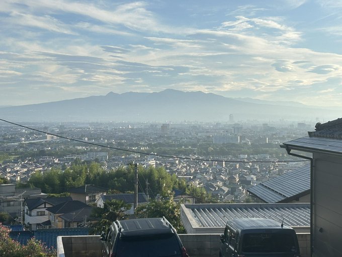 The image shows a scenic view of Akagi Mountain, visible in the distance under a clear sky with scattered clouds, as seen from a residential area. The foreground features rooftops, parked cars, and power lines, with a sprawling cityscape below the mountain. The post text, dated August 18 at 6:04 AM, mentions the end of the Obon festival and encourages a balanced approach to work amidst the summer heat, providing context that this is a morning view of the mountain in Gunma Prefecture. No platform watermarks are present.