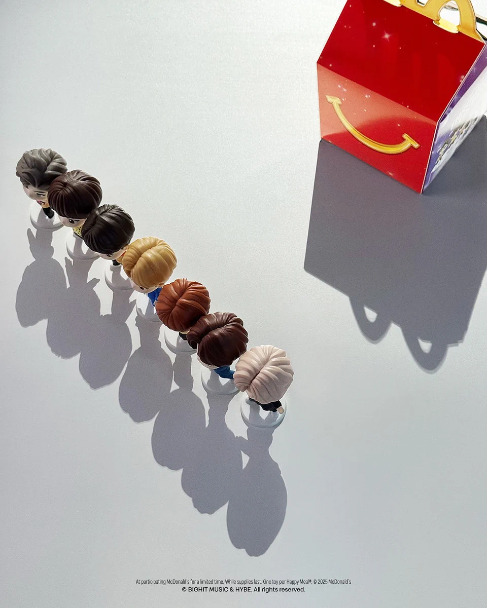 A 4:5 image showing a red Happy Meal box with hints of stars. The box is in the top right corner. In front of the box, a line made of 7 different little figurines side by side, seen from above, letting appear their glorious and magnificent hair style and colors. The image is lit in a way that casts a shadow on every single figurine and the box, adding dramatization to the iconic nature of the image itself. It is not possible to know or see who are represented on the 7 figurines, except if you have immaculate taste in music and human beings.