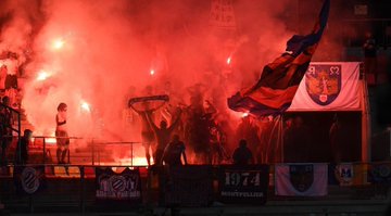 The image depicts an energetic scene of Montpellier Ultras fans at a Ligue 2 football match on August 19, 2025, between LMFC and MHSC, as indicated by the post text from @MHSC_ULTRAS. Fans are holding flares and a large flag with red, blue, and white colors, surrounded by red smoke, creating a vibrant stadium atmosphere. Banners display "Butte Paillade 1991" and "1974," referencing the fan group and its history, while the Montpellier crest is visible. The composition is dynamic, with fans cheering passionately, illuminated by the glow of flares, capturing the intensity of the event.