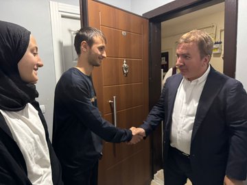 Hamza Aydoğdu in a suit shaking hands with Süleyman Adaş, who is sitting on a couch in a living room. Ufuk Kuyrukluyıldız and Hacı Ömer Kartal are present, along with Yunus Emre Adaş and other family members. The room has a gray couch, patterned rug, wooden door, and chairs.