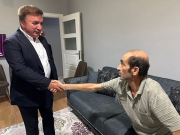 Hamza Aydoğdu in a suit shaking hands with Süleyman Adaş, who is sitting on a couch in a living room. Ufuk Kuyrukluyıldız and Hacı Ömer Kartal are present, along with Yunus Emre Adaş and other family members. The room has a gray couch, patterned rug, wooden door, and chairs.