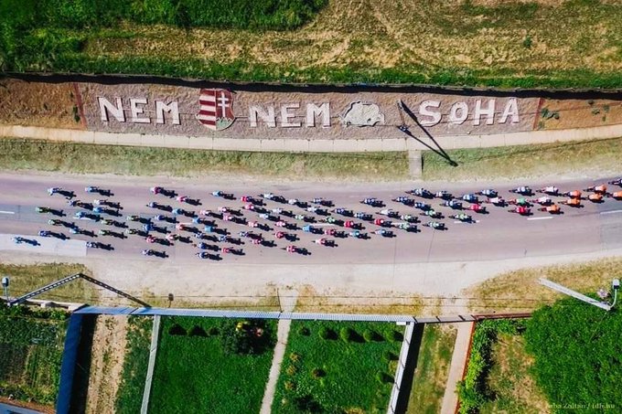 Cyclists riding on a road, forming a long line. Large text on a hillside reads "NEM NEM SOHA" with a coat of arms featuring red, white, and green colors. Green fields and a pathway are visible on either side of the road.