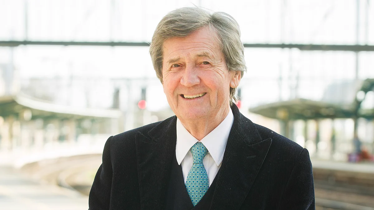 Melvyn Bragg smiles to camera wearing a dark suit jacket, white shirt and blue/green patterned tie. He stands at a railway station on the platform.