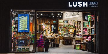 A black background with bold text reading "STOP STARVING GAZA" in red and green letters, resembling the Palestine flag colors, and smaller white text below stating "WE ARE CLOSED IN SOLIDARITY." A photo of a Lush store interior with shelves of skincare products, a sign reading "LUSH FRESH HANDMADE SKINCARE COSMETICS," and a person inside. A waving Palestine flag with red, black, white, and green horizontal stripes and a red triangle on the left side.