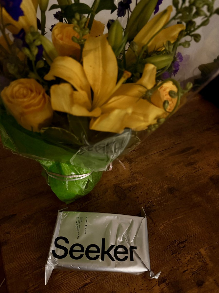 나무 테이블 위에 놓인 노란색과 보라색 꽃다발 옆에 있는 은색 Seeker 폰 패키지