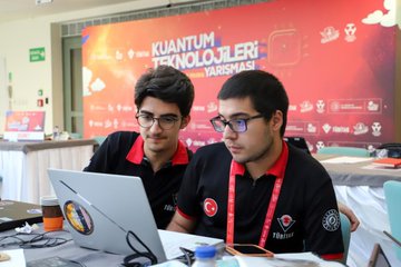 Orhan Aydın wearing a red jacket with arms crossed, talking to young men in black shirts with red collars and lanyards, standing indoors. Young men in black shirts with red collars and lanyards working on laptops at tables, indoors with a red backdrop displaying "Kuantum Teknolojileri Yarışması." A group of people, including Orhan Aydın in a red jacket, standing in front of a building with a sign reading "TÜBİTAK Ulusal Metroloji Enstitüsü."