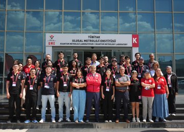 Orhan Aydın wearing a red jacket with arms crossed, talking to young men in black shirts with red collars and lanyards, standing indoors. Young men in black shirts with red collars and lanyards working on laptops at tables, indoors with a red backdrop displaying "Kuantum Teknolojileri Yarışması." A group of people, including Orhan Aydın in a red jacket, standing in front of a building with a sign reading "TÜBİTAK Ulusal Metroloji Enstitüsü."