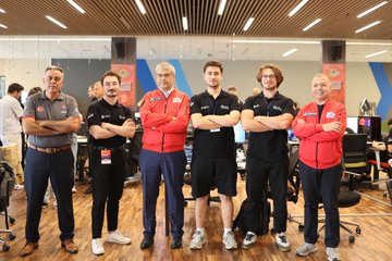 A group of people wearing red and black jackets with logos, standing and interacting in a modern indoor setting. Some individuals are gathered around a laptop displaying a screen with technical visuals. A banner in the background reads "Kuantum Teknolojileri Yarışması" with additional text and logos. Other people are posing together in a group photo, some in red jackets and others in black, in front of a banner with "13. ETÜCKS" and "Kuantum Teknolojileri Yarışması".