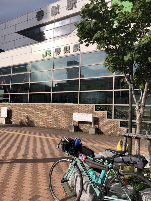 The image shows a bicycle parked in front of JR Kotoni Station (JR琴似駅) in Sapporo, Japan. The bicycle is equipped with various gear, indicating a long-distance cycling trip. The station building is modern with large glass windows and the JR logo prominently displayed. The post text indicates that the user has reached this station as their goal, celebrating with "やった〜🙌" (Yatta~! 🙌), which means "I did it!" in Japanese. The context from the original post text suggests that the user was on a challenging cycling route with rough roads, expressing a desire for better pavement.