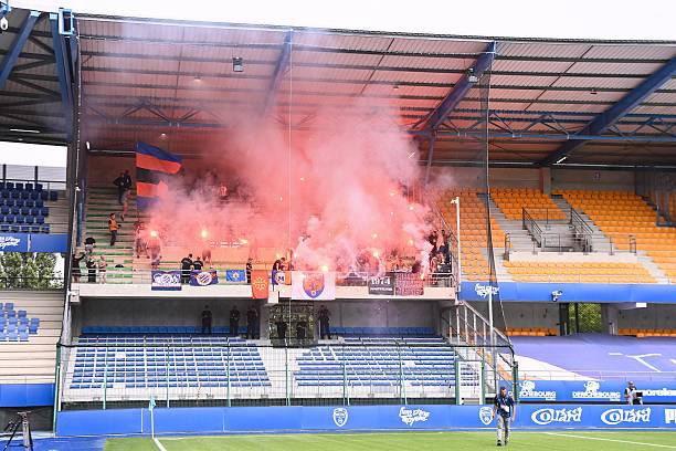 A stadium stand filled with fans, including Montpellier Ultras, holding red smoke flares. The stands have blue and orange seats, with banners and flags visible. The field is partially seen in the foreground.