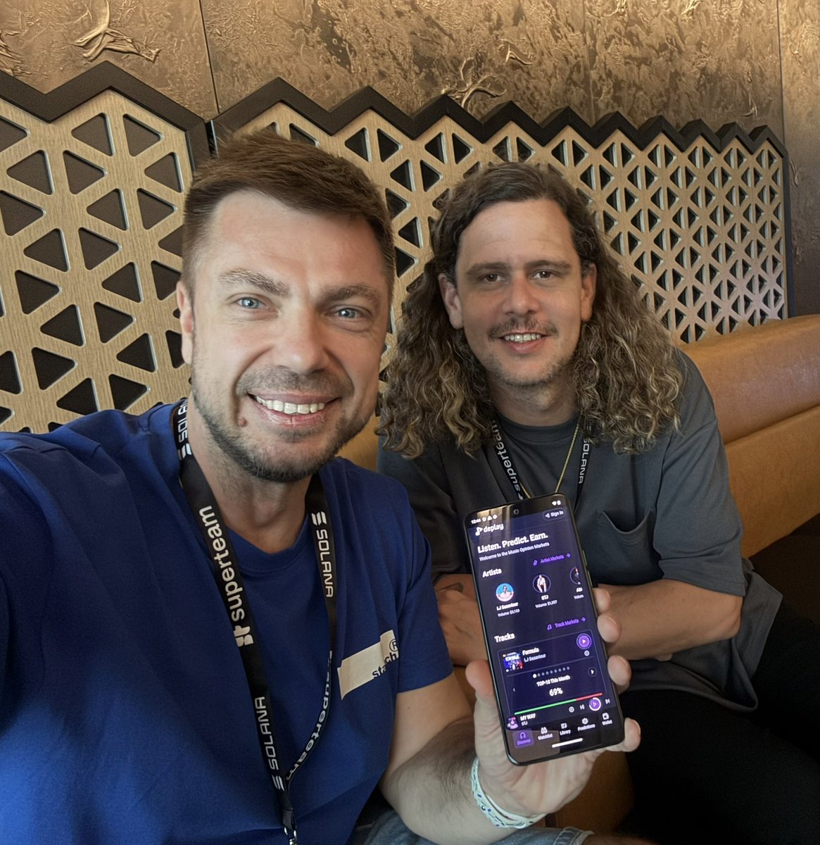 Serj Korj and Chase Barker smiling while holding a phone displaying the Deplay app at a Solana event