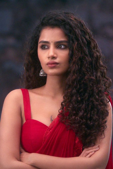 Anupama Parameswaran wearing a yellow saree with intricate patterns, adorned with gold jewelry including earrings and a necklace. Her hair is styled in loose curls, cascading over her shoulders. In another image, Anupama Parameswaran is dressed in a red saree with a sleeveless blouse, her arms crossed, and her curly hair flowing freely.