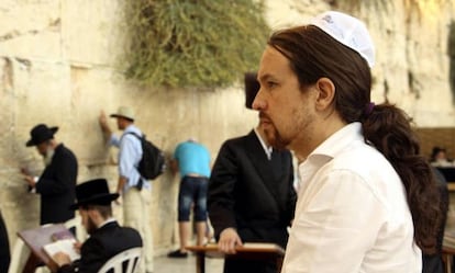 A man with long hair in a ponytail and a white kippah sits at a table, wearing a white shirt. Other individuals, some wearing traditional black hats and prayer shawls, stand or sit near a stone wall with greenery growing on it. The setting includes people engaged in prayer or reflection.