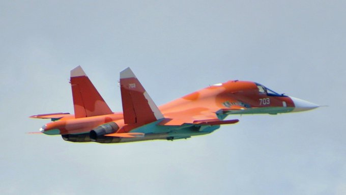 A Sukhoi Su-34 fighter-bomber in flight. The aircraft is painted orange with the number 703 visible on its fuselage. The sky is clear in the background.