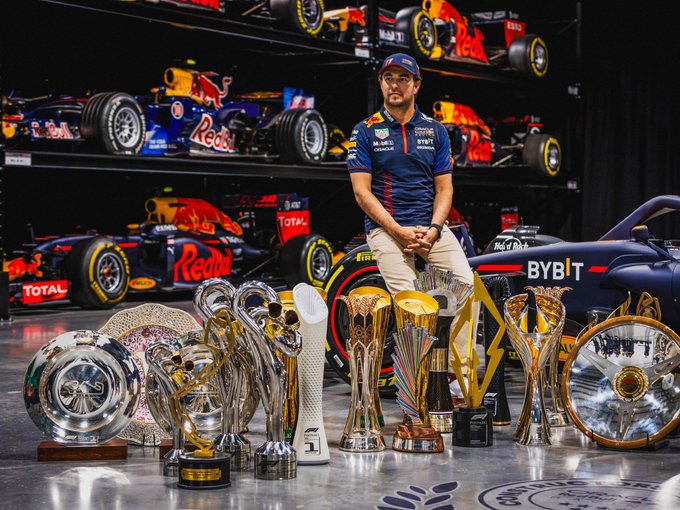 Sergio Pérez se para frente a una exhibición de autos de carreras de Fórmula 1 con la marca Red Bull y Cadillac. Lleva un traje de carreras azul marino y rojo. Frente a él hay varios trofeos y premios, incluidas grandes copas de plata y oro.