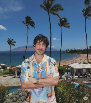 Shows Pietro Schirano wearing a colorful Versace shirt with AI clothing change effect, featuring floral and classical art patterns, with tropical beach resort background