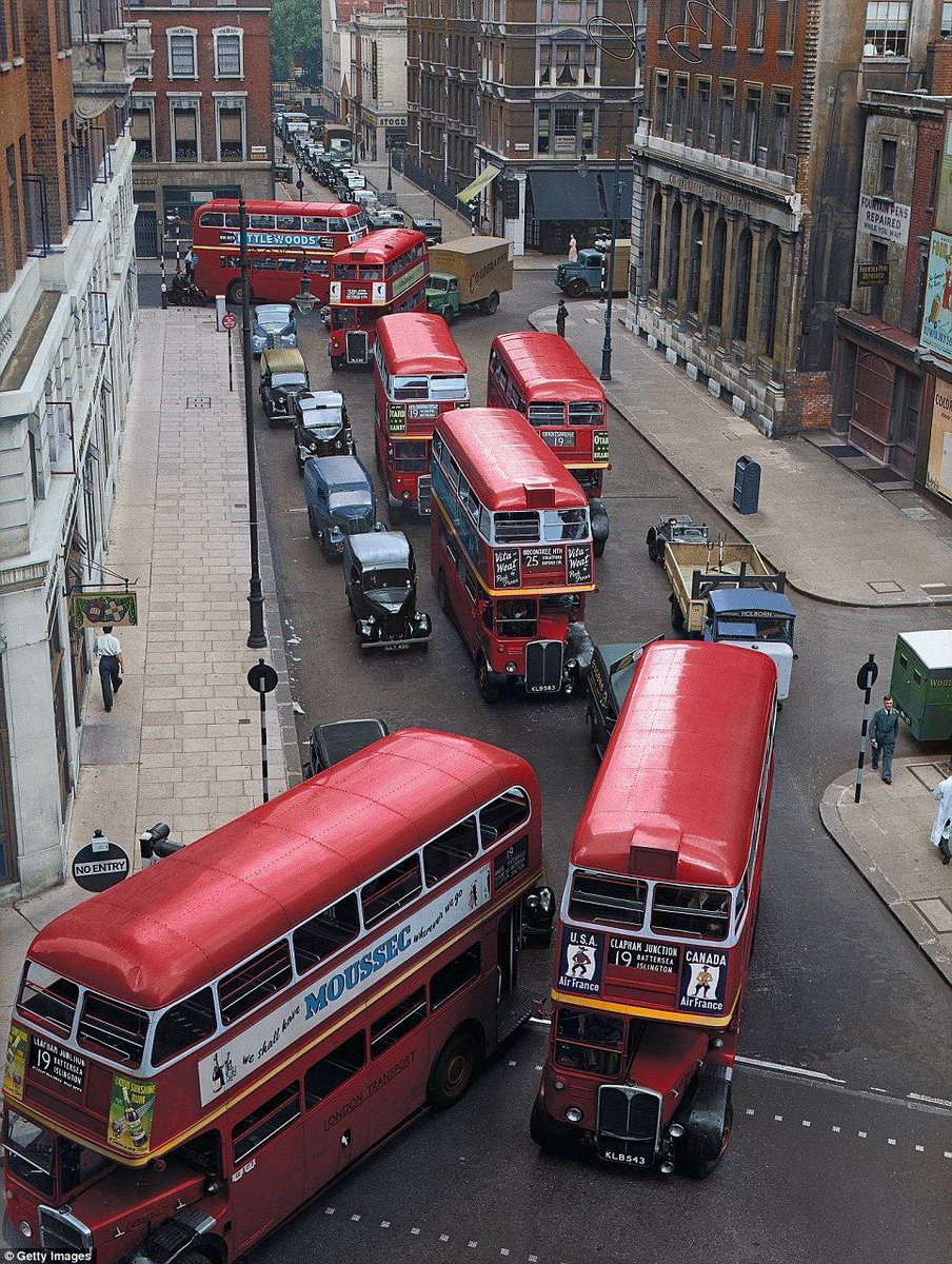 A photo from 1952 at Bury Place, London. A traffic jam of double decker buses and vintage cars next to a ‘Fountain Pens Repaired While You Wait’ sign.