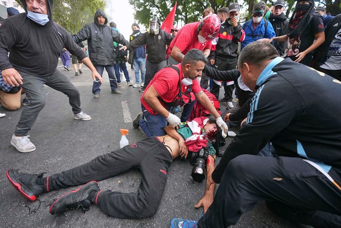 Several people in casual and athletic clothing, some wearing red shirts, attending to a person lying on the ground. The person on the ground has visible injuries, including blood on their legs and head. Others stand or kneel around, some gesturing actively, on a street with trees and a crowd in the background.