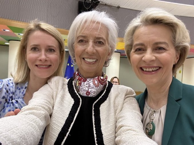 Three women smiling at the camera. Kaja Kallas, with blonde hair, wears a green blazer and necklace. The woman in the center, with white hair, wears a white jacket with black trim and a patterned scarf. The woman on the left, with blonde hair, wears a blue and white patterned blouse. European Union flags are visible in the background.