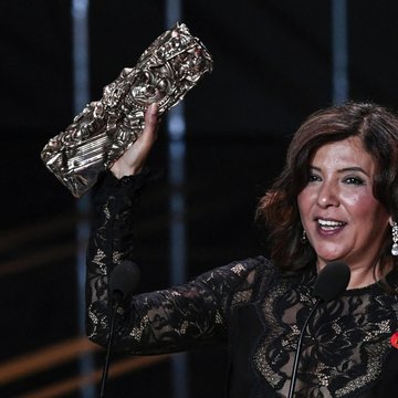 Three people, including a man in a white sweater holding a smartphone and two women in light-colored clothing, gathered around a laptop in a bright room. A woman in a black lace dress holding a golden award statue on stage with two microphones. A child in a black graduation gown and cap with Arabic text on the sash, holding the cap in one hand, standing indoors near a vase of tulips and a gold table.