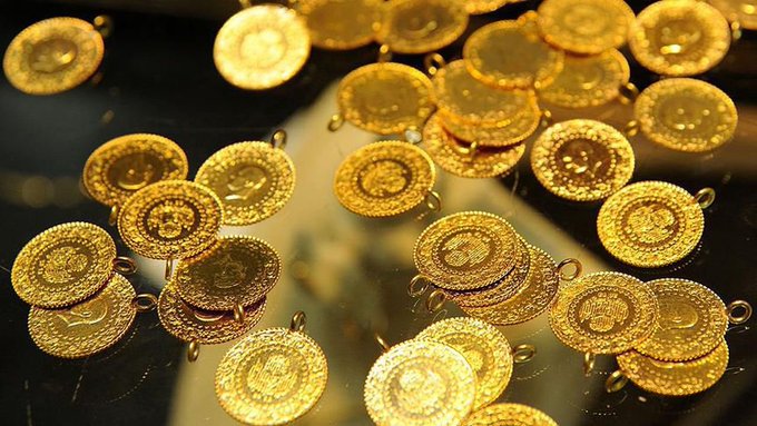 Multiple gold coins with intricate designs scattered on a dark surface. The coins are round and shiny, reflecting light, with visible engravings and patterns.