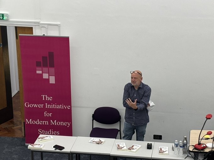 Warren Mosler standing at a table with copies of his book "Under the Bonnet" by Phil Armstrong. A banner for The Gower Initiative for Modern Money Studies is visible behind him. Phil Armstrong and Carlos Garcia stand nearby, with tables holding water bottles, microphones, and papers. The setting appears to be a conference room.