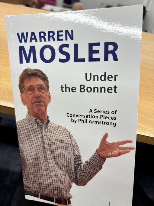 Warren Mosler standing at a table with copies of his book "Under the Bonnet" by Phil Armstrong. A banner for The Gower Initiative for Modern Money Studies is visible behind him. Phil Armstrong and Carlos Garcia stand nearby, with tables holding water bottles, microphones, and papers. The setting appears to be a conference room.