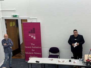 Warren Mosler standing at a table with copies of his book "Under the Bonnet" by Phil Armstrong. A banner for The Gower Initiative for Modern Money Studies is visible behind him. Phil Armstrong and Carlos Garcia stand nearby, with tables holding water bottles, microphones, and papers. The setting appears to be a conference room.