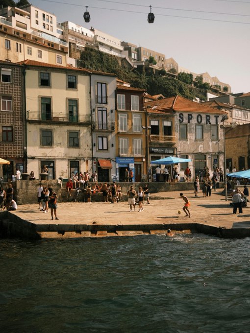 A waterfront scene in Porto, Portugal, with colorful buildings lining the shore. People are gathered on a stone platform by the water, some playing or walking. A cable car is visible in the sky, and a building with the word "SPORT" is prominent. The water reflects the buildings and people along the edge.