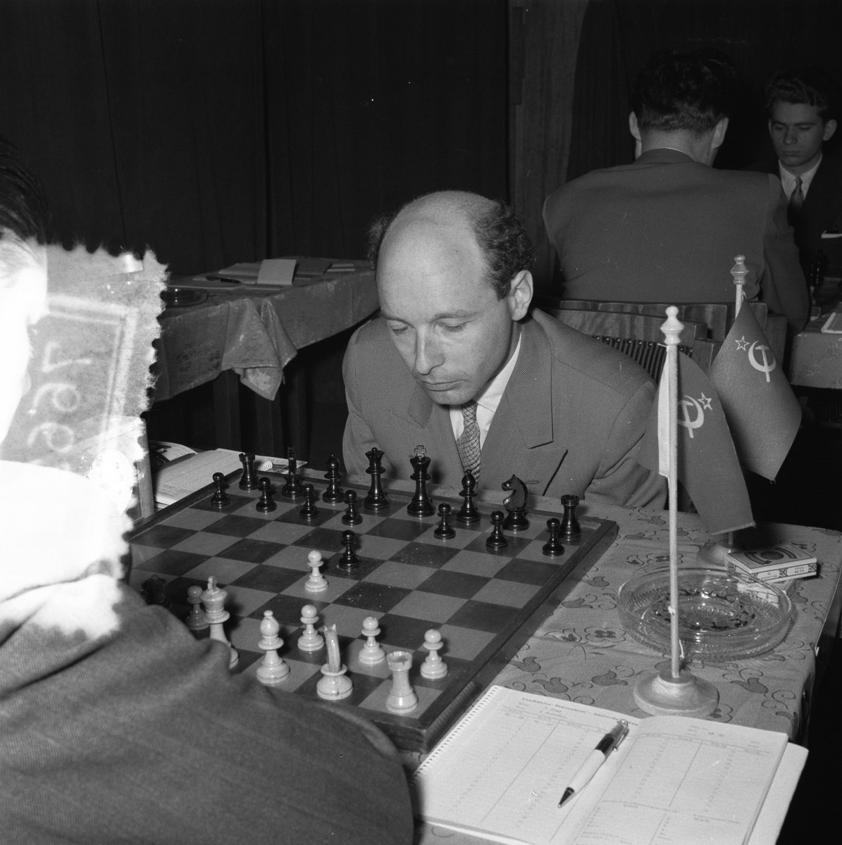 dgriffinchess's tweet image. David Bronstein, pictured in play v. Paul Keres in the opening round of the 1956 Candidates' tournament in Amsterdam. In the background, the young Boris Spassky is seen in play v. Miroslav Filip.
(📷: H. Behrens / ANEFO, via nationaalarchief.nl.) #chess