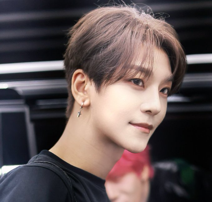 He Xinlong wearing a black mask, black shirt, and black jacket. He has short dark hair, a cross earring, and a necklace. A watermark reading "Graced" is visible.