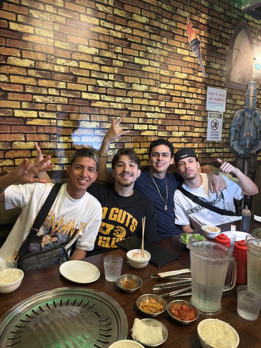 Four people are sitting at a table in a restaurant with a brick wall background. They are posing for the photo with various hand gestures. The table is filled with food items including a bowl of soup, a pitcher of water, a bottle of ketchup, and various small dishes with sauces and side items. One person is wearing a white shirt with a graphic design, another is wearing a black shirt with yellow text, and another is wearing a white shirt with a black strap across the chest. There is a statue of a knight and a torch on the wall behind them.
