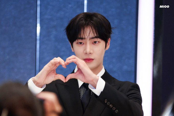 Choo Youngwoo is seen wearing a black suit with a white shirt and black tie. He is making a heart gesture with his hands. The background is colorful with a mix of red and blue lights. There are people partially visible in the foreground, suggesting a public or fan event setting. The image has a watermark in the top right corner that reads "MIOO".