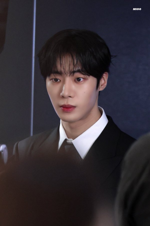 A young man with black hair wearing a black suit and white shirt. A watermark reading "mioo" is visible in the top right corner.