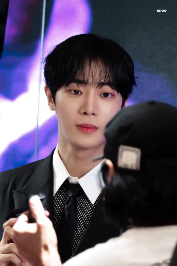 Choo Youngwoo wearing a black suit, white shirt, and black tie, standing in front of a purple and black background. He has black hair and is holding a small object in his hand. Another person wearing a black cap is partially visible, adjusting or interacting with Choo Youngwoo\'s appearance.