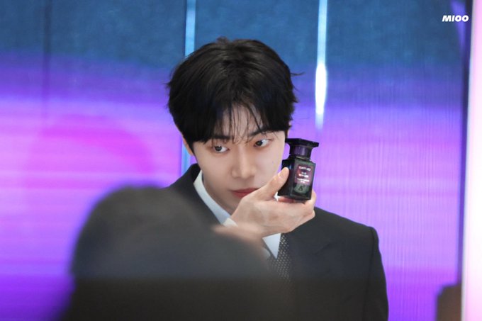 Miwoo wearing a black suit and tie, holding a small black bottle in his right hand. He has short black hair and is looking downward. A person in the foreground partially obscures the view, and a vibrant purple and blue background with vertical lines is visible. A watermark reading "MIOO" is present in the top right corner.