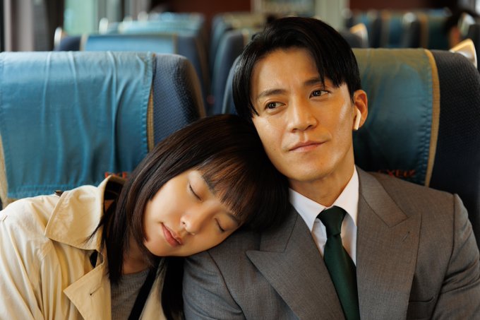 Shun Oguri and Han Hyo-joo sitting on a train. Shun Oguri, wearing a gray suit and green tie, sits upright. Han Hyo-joo, wearing a beige coat, rests her head on Shun Oguri\'s shoulder, appearing to sleep. Blue train seats are visible in the background.