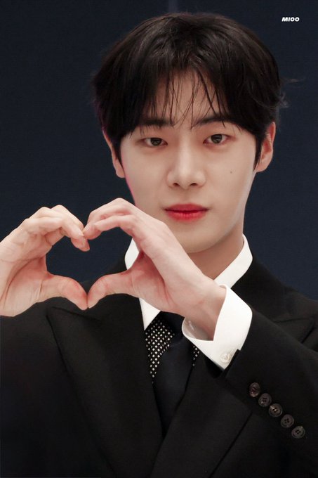 Choo Youngwoo is wearing a black suit with a white shirt and a black tie with white polka dots. He is making a heart shape with his hands. The background is dark, likely a stage or event setting.