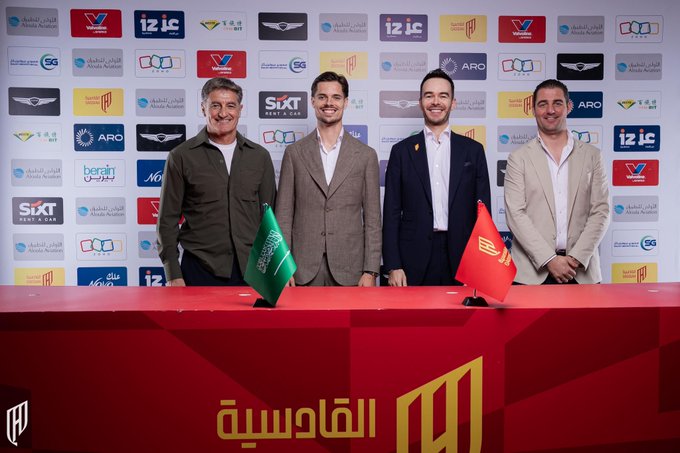 Four men are standing behind a table with two flags, one green and one red, placed on it. The backdrop features various logos of sponsors and organizations. The men are dressed in formal attire, with one wearing a green jacket, another in a brown suit, another in a dark suit, and the last in a light-colored suit. The text on the table reads "القادسية" with a logo.