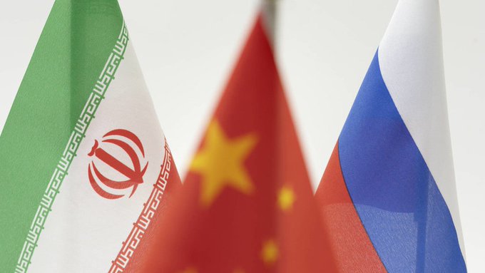 Tres banderas nacionales se exhiben de manera prominente. De izquierda a derecha, son las banderas de Irán, China y Rusia. La bandera iraní presenta rayas horizontales verdes, blancas y rojas con el emblema nacional en rojo. La bandera china tiene una gran estrella amarilla con cuatro estrellas más pequeñas en un semicírculo sobre un fondo rojo. La bandera rusa consta de tres franjas horizontales de blanco, azul y rojo.