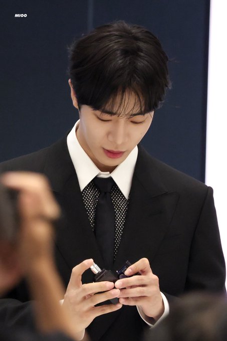 Choo Youngwoo is seen wearing a black suit with a white shirt and a black tie with white polka dots. He is holding a small black object in his hands, possibly a phone or a device, while looking down at it. There are people around him, one of whom is partially visible in the foreground, also holding a phone. The background is dark with some light reflections.
