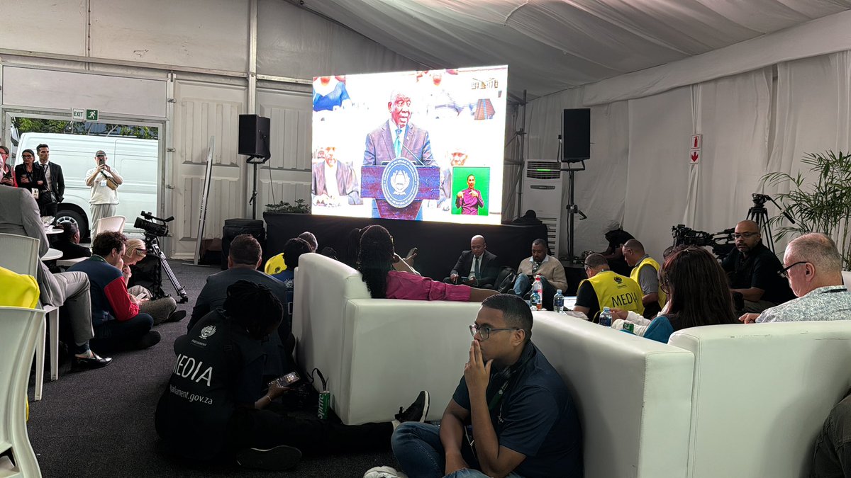 Journalists and media crew seated on couches and floor in a tented viewing area watching large screen of President Cyril Ramaphosa speaking during the 2026 State of the Nation Address broadcast.