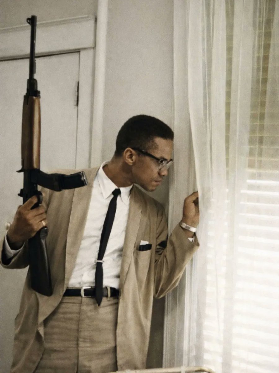 Malcolm X looking out the window holding a rifle, Queens, New York, 1964.