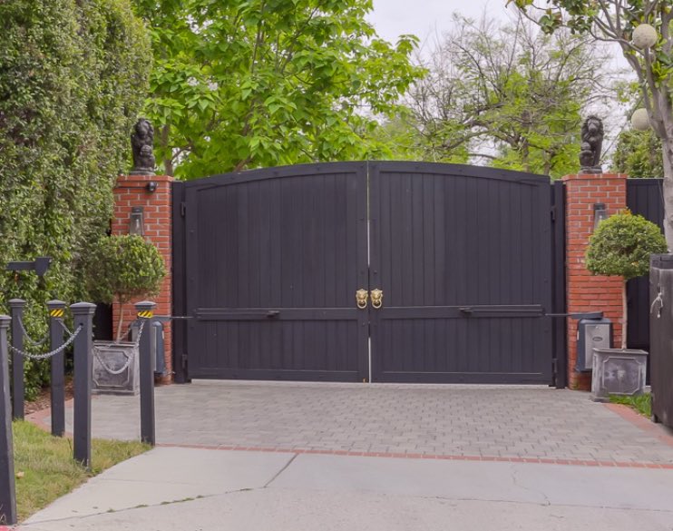 Luxurious black double gate with gold lion-head knockers, flanked by brick pillars topped with lion statues, surrounded by lush green trees and security features like bollards and chains.
