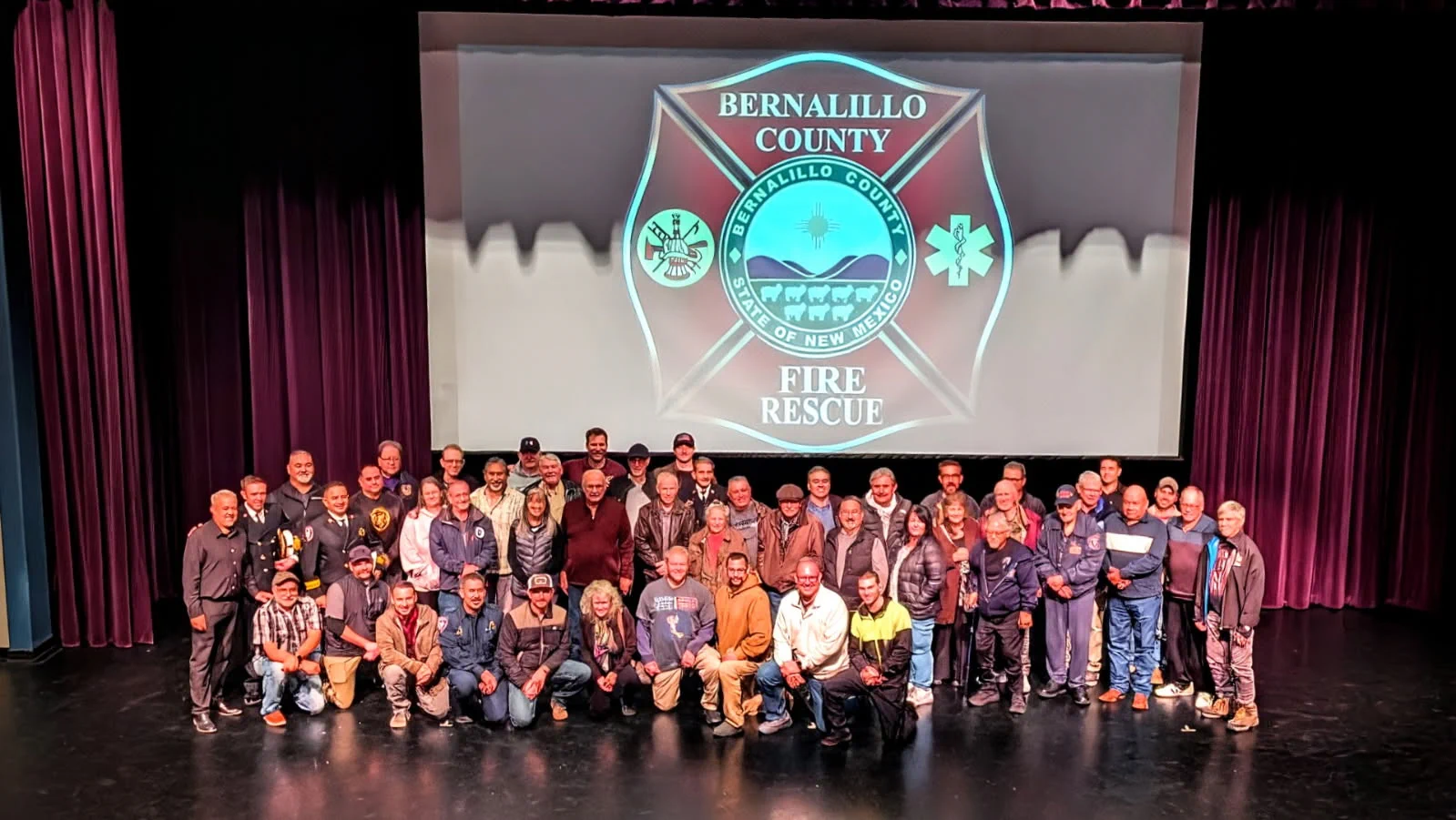 A group of people is gathered onstage in a theater, posing for a photo. Behind them is a large screen displaying the logo for Bernalillo County Fire Rescue. The stage curtains are visible on the sides.