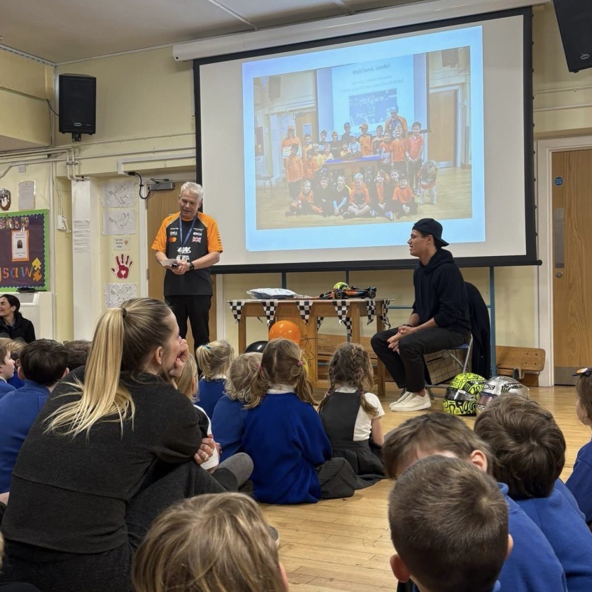 autosport's tweet photo. Lando Norris paid a very special visit to his old primary school in Bristol, where he took part in a whole-school assembly and spoke to kids about the importance of perseverance 🧡

The F1 World Champion visited every classroom, answered questions, signed items, and even waved the chequered flag for a schoolyard scooter race 🏁

He made sure every pupil received a gift before he left 🎁
