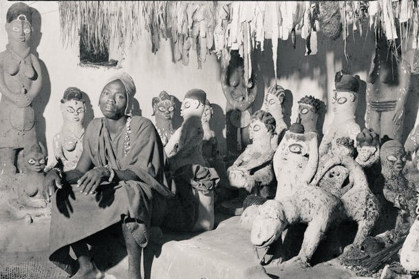 My kulture is not demonic....
A priest among gods

A man sitting among many figures dedicated to different divinity and deities, Benin city, ca.1950s.