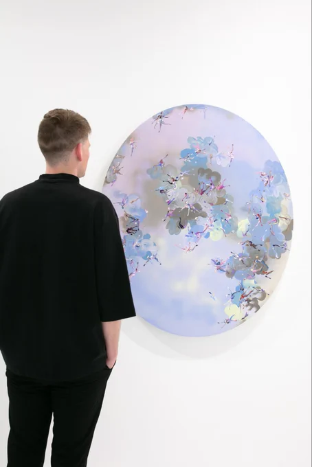 Installation view of Paul Kuntze solo exhibition “Falling Angels” with a guest standing in front of an oval shaped painting featuring gradient, glowing clouds, abstracted figures, and lush swirl brush strokes. The hues are blues, pinks, lavenders, and light pinks.