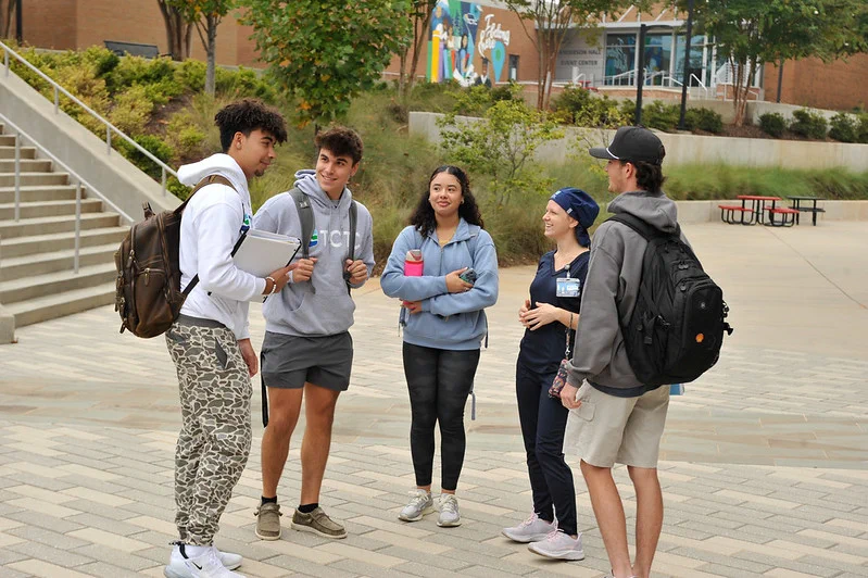 A group of five people stand outside, talking and smiling. They are in a paved area with stairs and trees nearby. Some hold backpacks or water bottles. The setting appears to be a college campus, with a mural visible in the background.