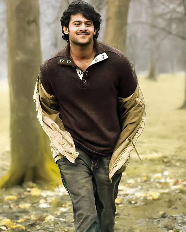 Create a hyper-realistic cinematic full-body portrait of a tall, muscular Indian man resembling Prabhas, walking casually toward the camera in a misty forest. He has medium-length wavy black hair, a light beard, and a warm confident smile. He is wearing a dark brown Henley shirt